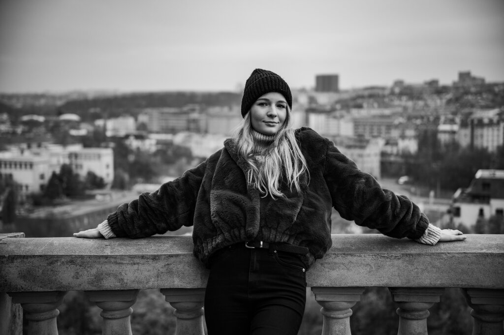 Černob&iacute;l&yacute; portr&eacute;t usměvav&eacute; blond&yacute;ny opřen&eacute; o z&aacute;bradl&iacute; s panoramatem Prahy.