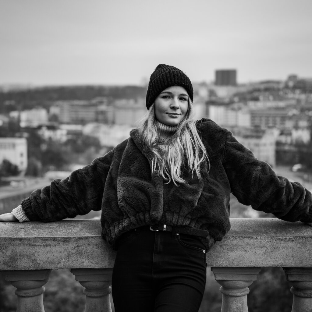 Černob&iacute;l&yacute; portr&eacute;t usměvav&eacute; blond&yacute;ny opřen&eacute; o z&aacute;bradl&iacute; s panoramatem Prahy.