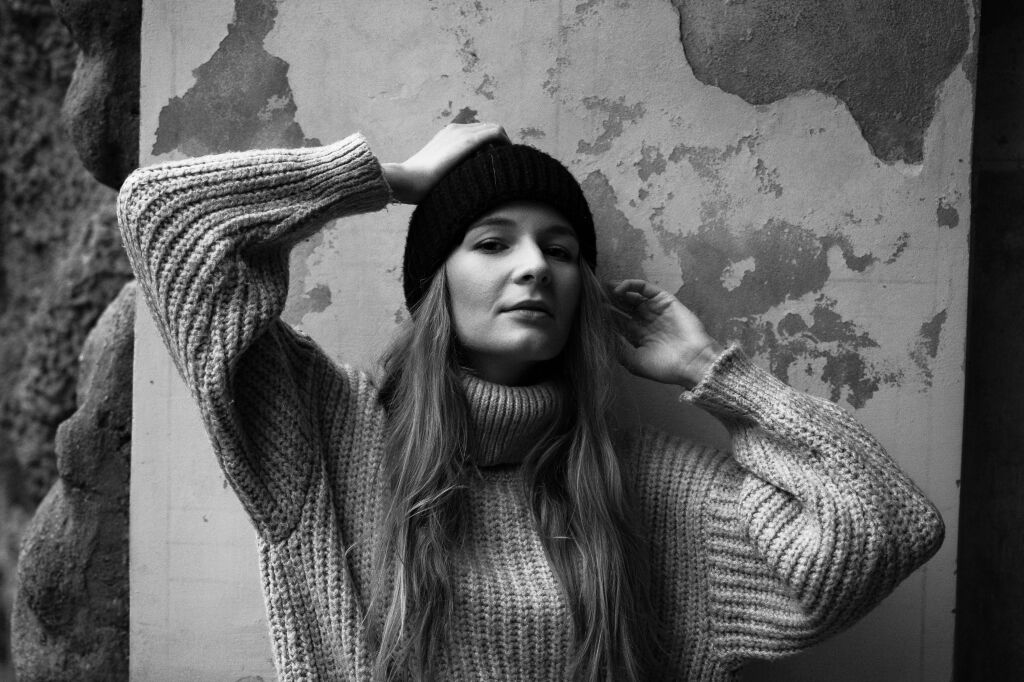 Černob&iacute;l&yacute; portr&eacute;t ženy v pleten&eacute;m svetru a čepici před starou zd&iacute; s oloupanou om&iacute;tkou.