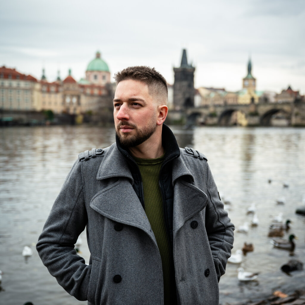 Portr&eacute;t muže v &scaron;ed&eacute;m kab&aacute;tě u Vltavy s Karlov&yacute;m mostem v pozad&iacute;.