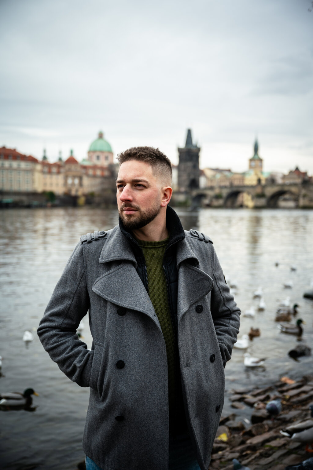 Portr&eacute;t muže v &scaron;ed&eacute;m kab&aacute;tě u Vltavy s Karlov&yacute;m mostem v pozad&iacute;.