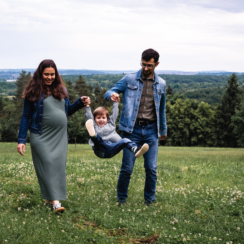 Těhotn&aacute; maminka s tat&iacute;nkem houpou synka za ruce na zelen&eacute; louce.