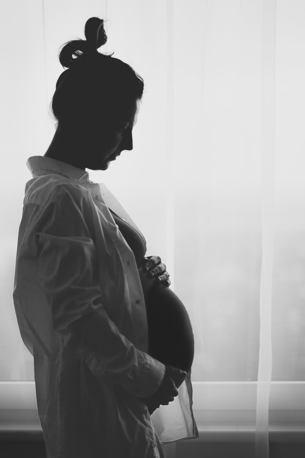 Černob&iacute;l&aacute; silueta těhotn&eacute; ženy u okna drž&iacute;c&iacute; si bř&iacute;&scaron;ko.