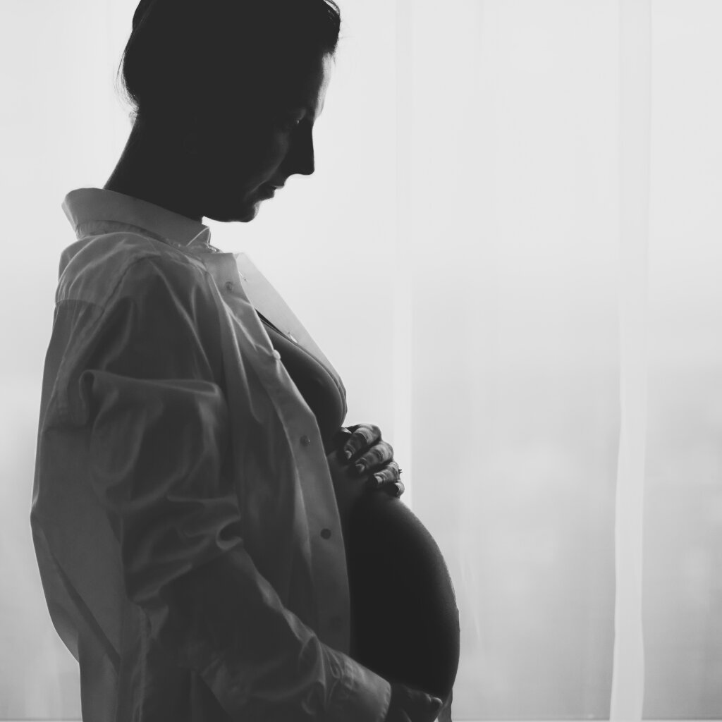 Černob&iacute;l&aacute; silueta těhotn&eacute; ženy u okna drž&iacute;c&iacute; si bř&iacute;&scaron;ko.