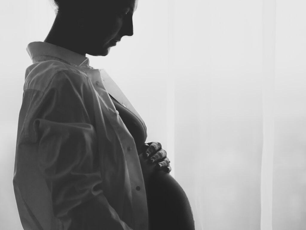 Černob&iacute;l&aacute; silueta těhotn&eacute; ženy u okna drž&iacute;c&iacute; si bř&iacute;&scaron;ko.