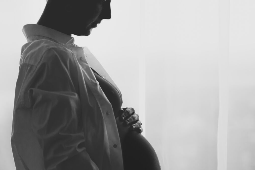 Černob&iacute;l&aacute; silueta těhotn&eacute; ženy u okna drž&iacute;c&iacute; si bř&iacute;&scaron;ko.